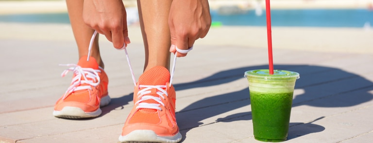 woman  on juice diet running tying shoelace with cup of fresh green juice next to her 