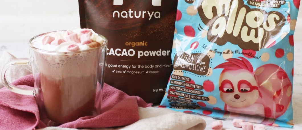 cacao powder hot chocolate in a mug next to Naturya cacao powder and bag of vegan marshmallows