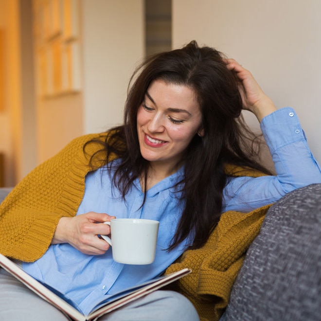 A photo of a woman smiling as she drinks a cup of tea under a blanket on a comfy sofa. She is probably listening to the Lord of the Rings audio book series.