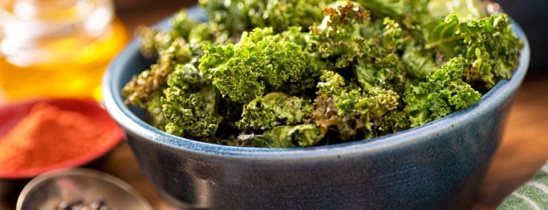 crispy kale in a bowl sat on a wooden table with oil, spices and peppercorn around it
