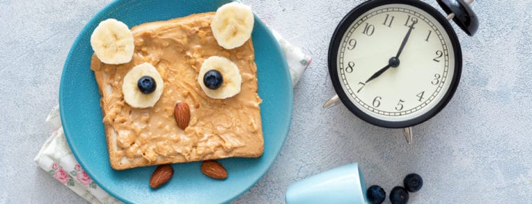 Funny owl toast with peanut butter, banana, blueberries and almonds on a blue plate, a clock and tiny cup of blueberries.