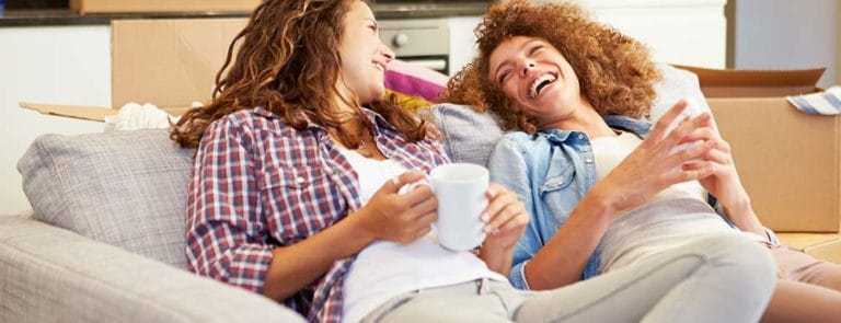 Two female friends sitting on a sofa drinking tea and laughing