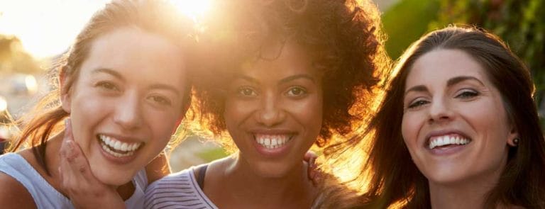 Three women with white teeth smiling at the camera with the sun behind them