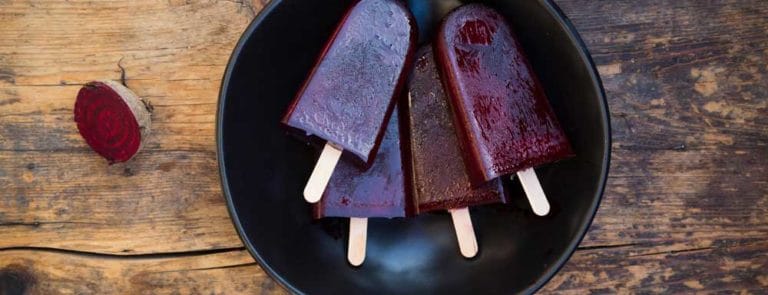 Purple ice lollies in a black bowl next to some halved beetroots on a wooden table