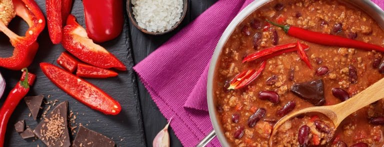 chili con carne with whole red hot chilis, kidney beans, tomatoes and piece of chocolate in a pot with ingredients at background, horizontal view from above, close-up