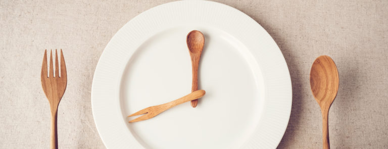 A plate with a spoon and fork resembeling the hands of a clock and two hands holding a fork in one hand and a spoon in the other.