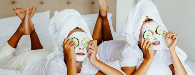 Two ladies in face masks, with cucmbers on their eyes, lying on a bed whilst wearing wrapped towels around their heads.