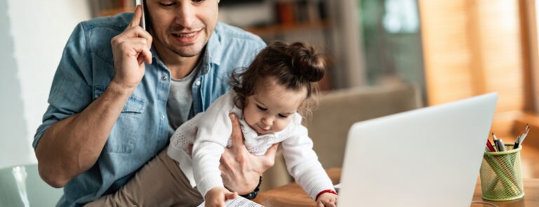 A man working from home whilst holding is baby daughter in one hand and trying to speak on the phone in the other hand.