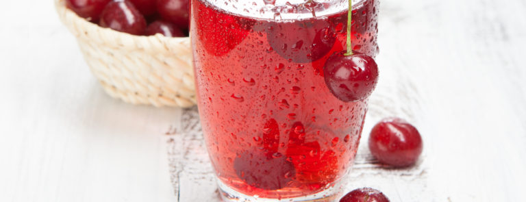 A bowl of cherries with a glass of cherry juice in front and some cherries scattered in the juice and on the table.