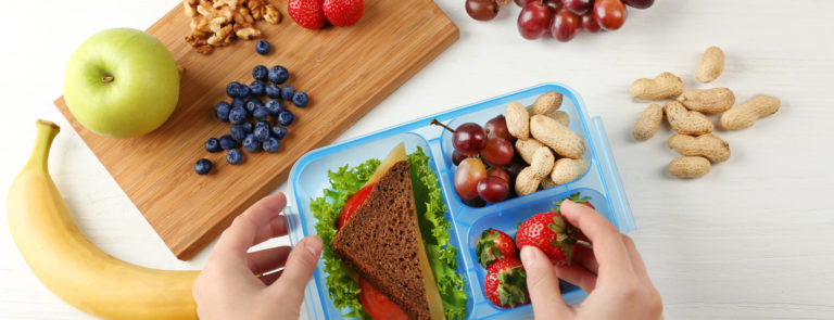 A lunch box with a sandwich and variety of fruits and nuts included. As well as a chopping board with nuts, blueberries, strawberries and grapes around it.