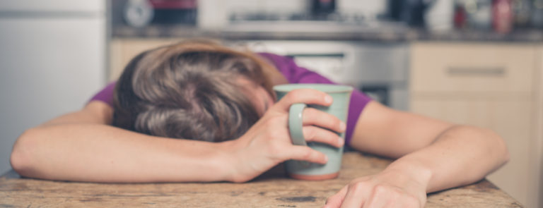 A lady asleep at her desk whilst holding a mug in one hand.