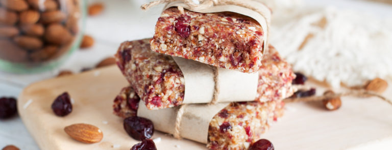 Three protein oat bars with cranberries and nuts in, piled on top of each other. They are on top of a chopping board with nuts and fruit scattered around.
