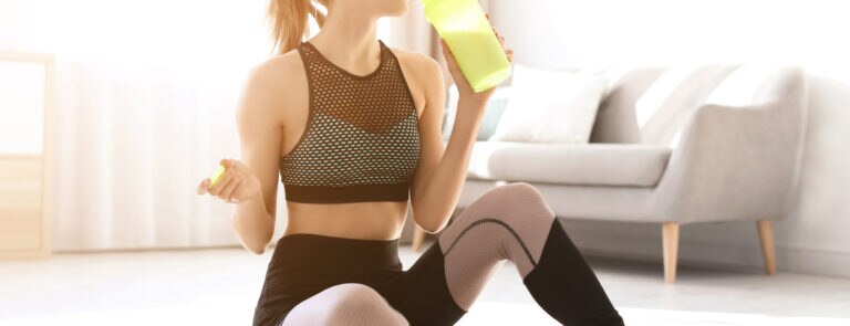 A lady sitting on her floor in sportswear, drinking a nutritious drink from a shaker cup.